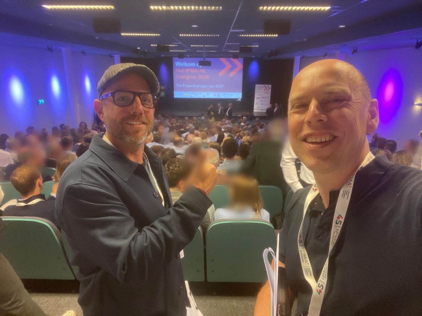 De volle congreszaal met enkele honderden deelnemers, gezien vanaf de zijkant, met het welkomstscherm op de achtergrond.