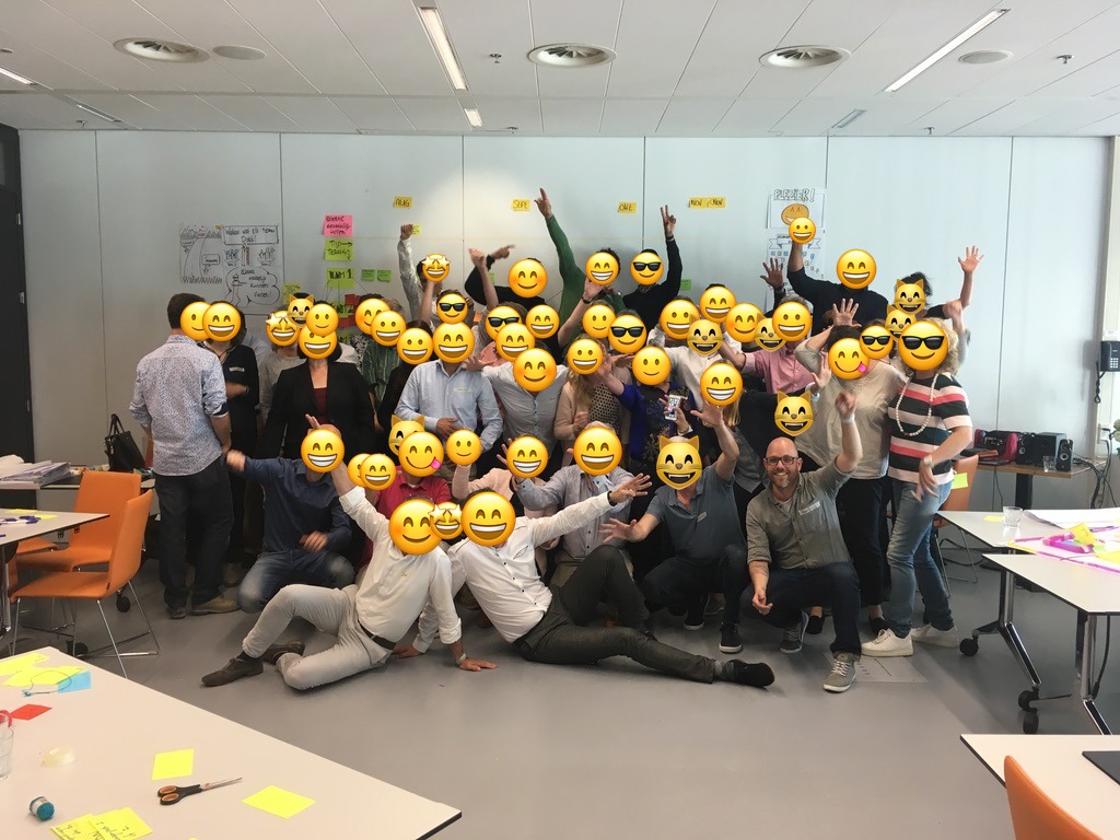 Een grote groep aan het eind van een werksessie, armen omhoog, in een werkruimte met handgetekende flipovers langs de wanden. De gezichten van de deelnemers zijn afgedekt met smileys.